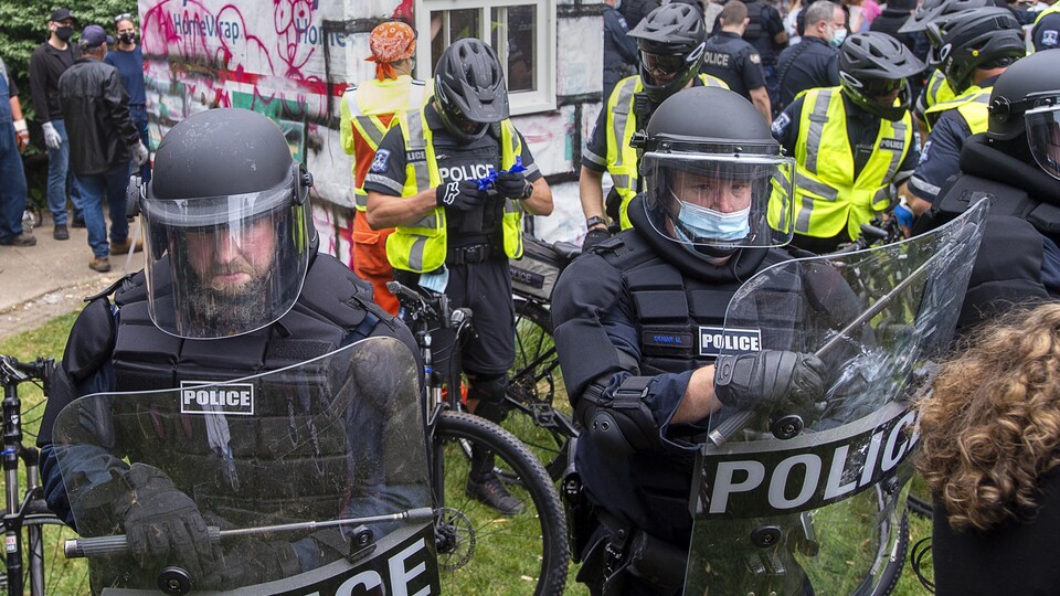 Une rangée de policiers en tenue antiémeute, avec des boucliers, devant des policiers en vélo autour d'une cabane habitée par une personne sans abri dans un parc.