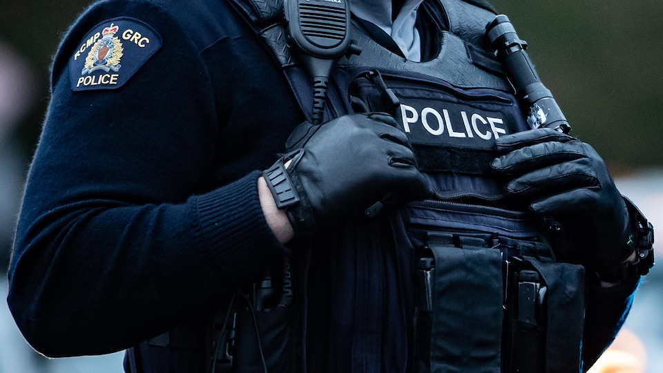 Plan rapproché de la poitrine d'un policier en uniforme couverte d'une veste noire décorée de l'emblème de la GRC, d'un walkie-talkie, d'une lampe de poche et d'un gilet pare-balles sur lequel le mot « Police » est écrit.