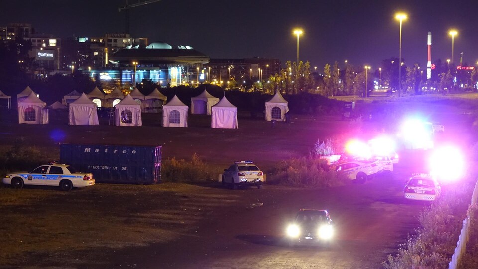 Des voitures de police, gyrophares allumés, entourent un site où sont plantées de petites tentes blanches, à Laval.