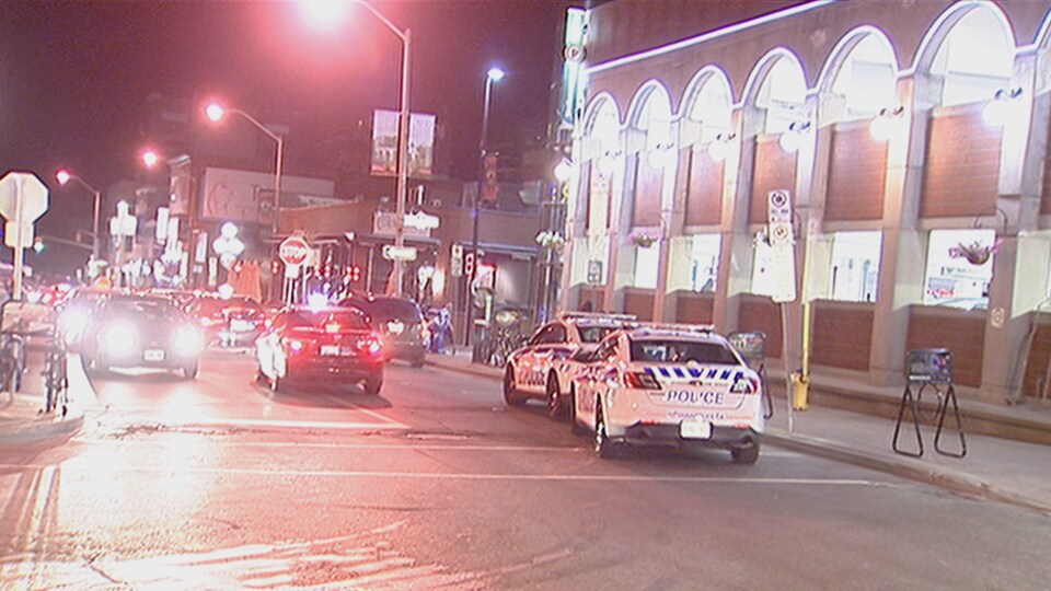 Deux voitures de police la nuit, dans une rue passante où plusieurs autres véhicules circulent.
