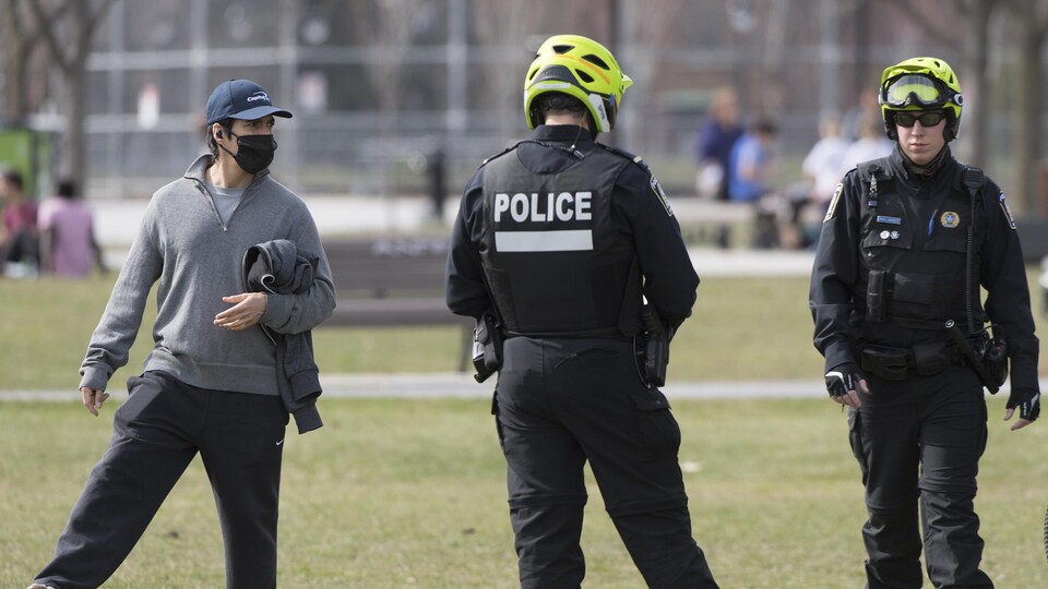 Deux policiers dans un parc près d'un homme portant un masque et posant le pied sur un ballon de soccer.