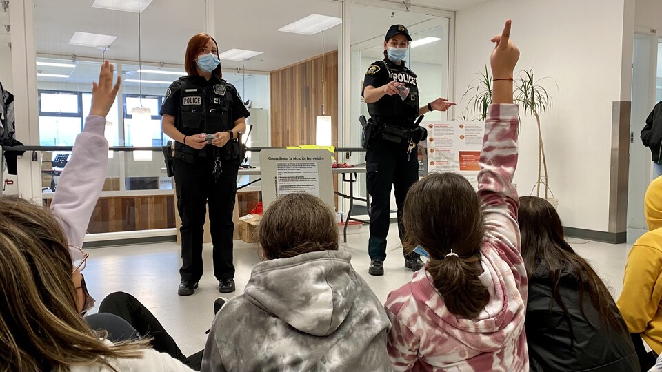Des élèves sont assis au premier plan et écoutent une discussion animée par deux policières dans une école.