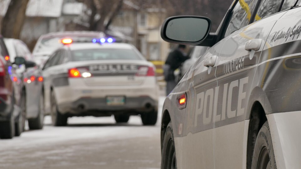 Gros plan sur deux voitures du Service de police de Winnipeg, l'une en avant de l'autre. Au loin, on voit la bande jaune qui délimite un périmètre de sécurité, et un agent.