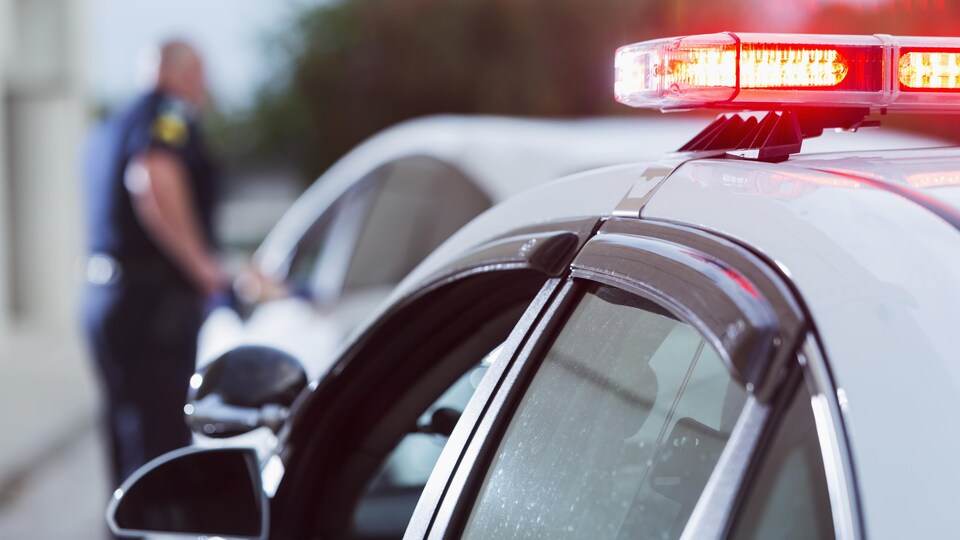 Une voiture de police arrêtée en bordure de la route. Derrière, flou, un policier en uniforme, debout à côté de la portière d'une automobile.