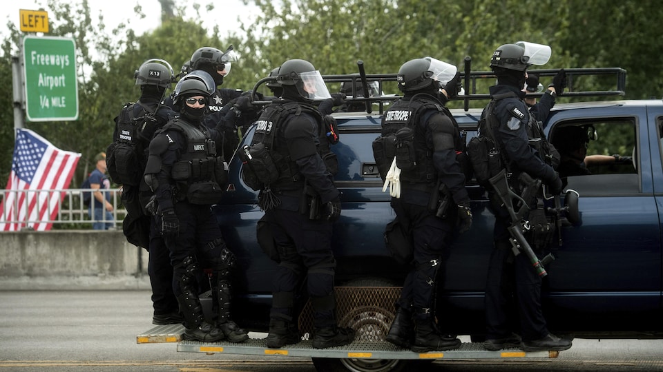 Des policiers armés debout sur une plateforme autour d'un véhicule qui roule sur la route.