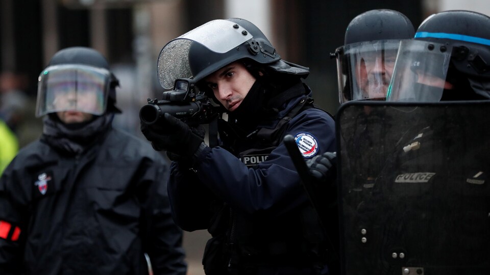 Un policier français en tenue antiémeute tient un fusil éclair alors que des manifestants prennent part à une manifestation du mouvement des « gilets jaunes » sur la place de la Bastille à Paris.