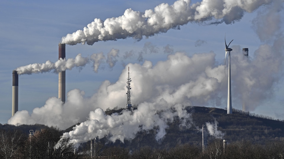 Une centrale alimentée au charbon et une raffinerie dégagent des volutes de fumée aux côtés d'une éolienne.