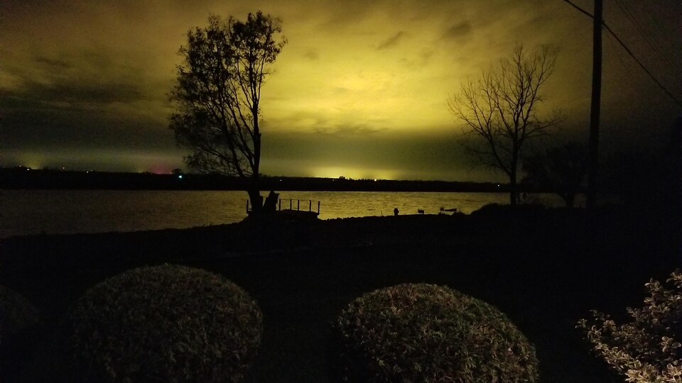 Vue de nuit d'une étendue d'eau depuis le terrain d'une résidence. Le ciel est jaune par endroits en raison de la lumière émise par des serres agricoles.