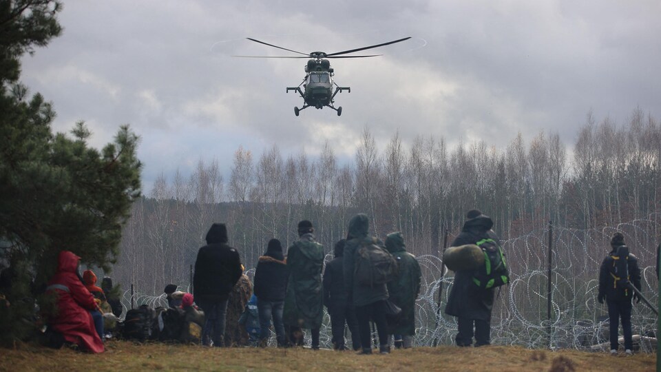 Des migrants massés devant des barbelés et un hélicoptère qui vole au-dessus d'eux.