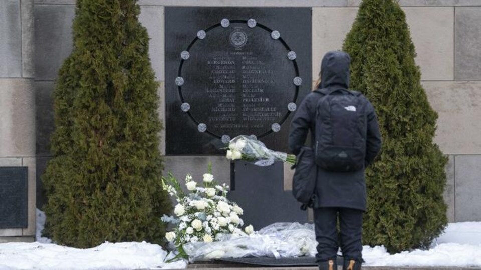 Une femme portant un capuchon tient une gerbe de roses blanches et se recueille devant le mémorial.