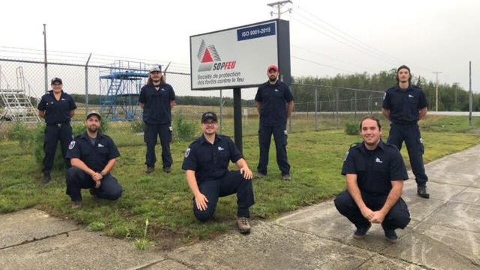 Sept pompiers posent chacun à deux mètres de distance devant une affiche de la SOPFEU.