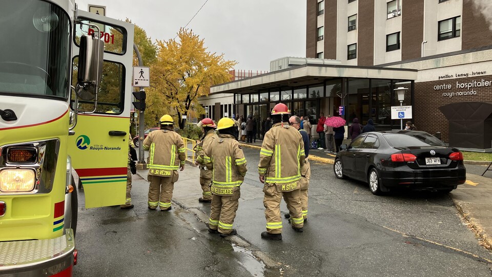 Six pompiers discutent devant l'Hôpital de Rouyn-Noranda. Une vingtaine de personnes attendent devant l'entrée.