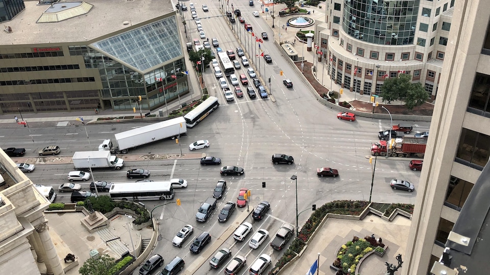 On voit de haut une intersection importante dans un centre-ville, avec des voitures et des camions qui circulent. On peut ausi voit qu'il n'y a pas d'endroits permettant aux piétons de traverser la rue sur cette intersection.