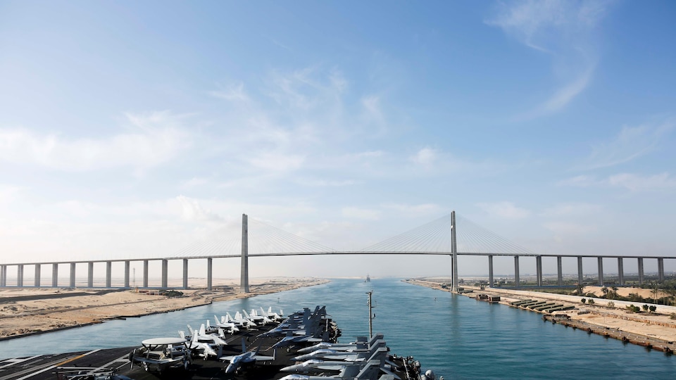 La proue du porte-avions s'apprête à franchir un pont au-dessus du canal de Suez. Des chasseurs sont stationnés sur le pont.