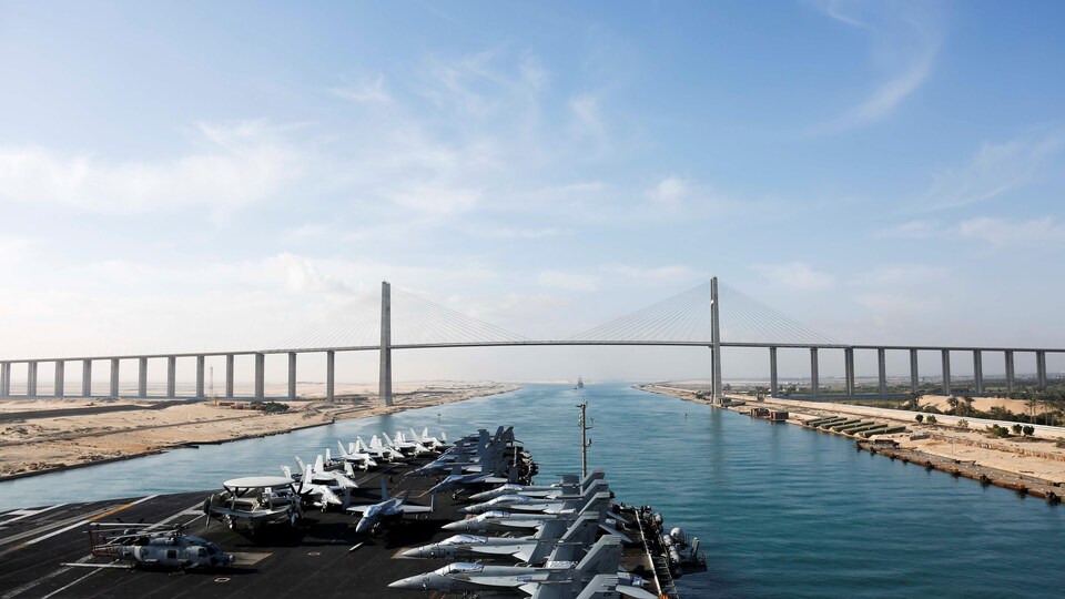 La proue du porte-avions s'apprête à franchir un pont au-dessus du canal de Suez. Des chasseurs sont stationnés sur le pont. 