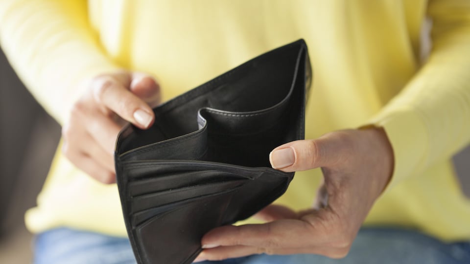 Une femme tient dans ses mains un portefeuille vide. Elle porte un pull jaune et un jean. Le portefeuille est en cuir noir.
