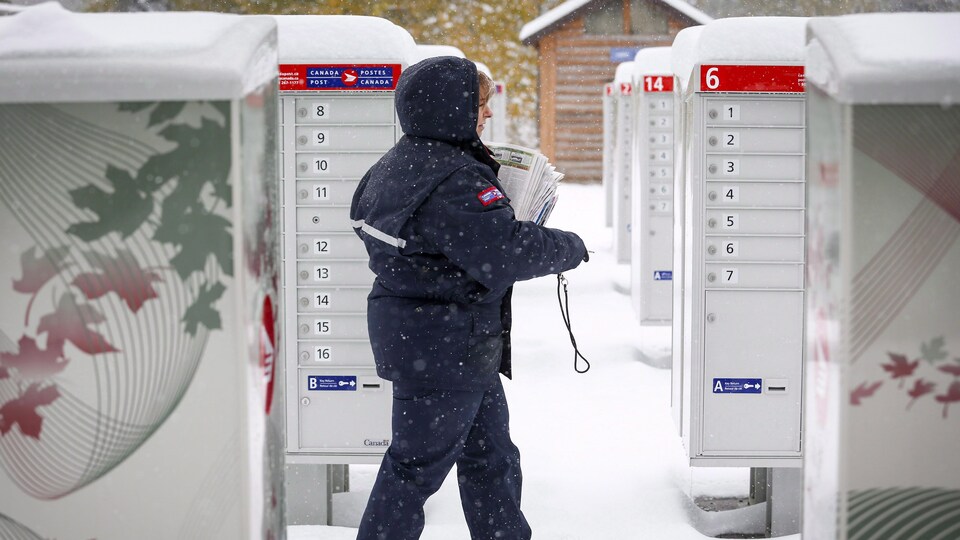 Une femme en vêtements d'hiver aux couleurs de Postes Canada marche dans la neige et s'approche de boîtes à lettres communautaires.
