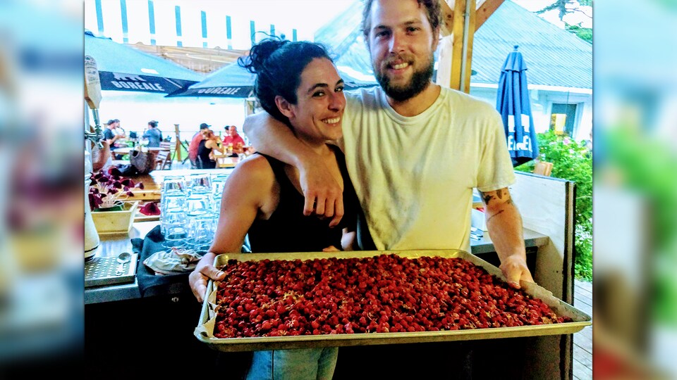 Dans un marché public, une femme et un homme sourient et tiennent un plateau de petites fraises. L'homme enlace la femme d'un bras.
