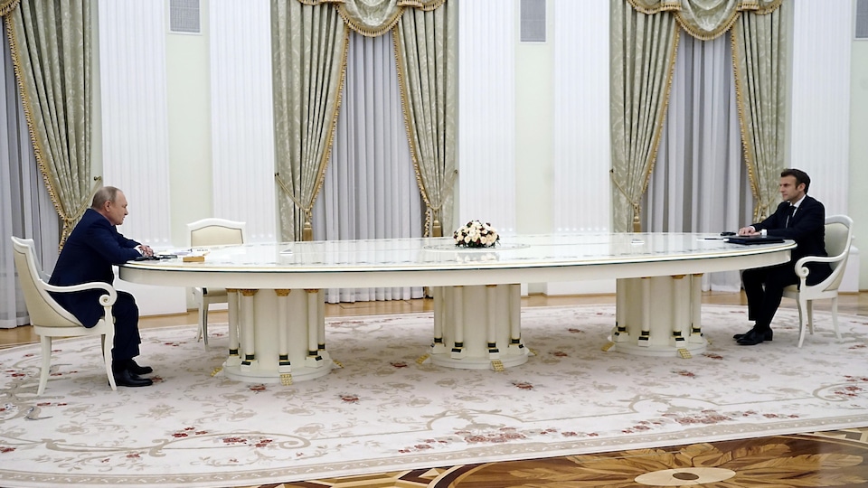 Vladimir Poutine (à gauche) est assis au bout d'une longue table blanche devant Emmanuel Macron (à droite). Ce somptueux décor est celui d'une salle du palais du Kremlin, à Moscou.