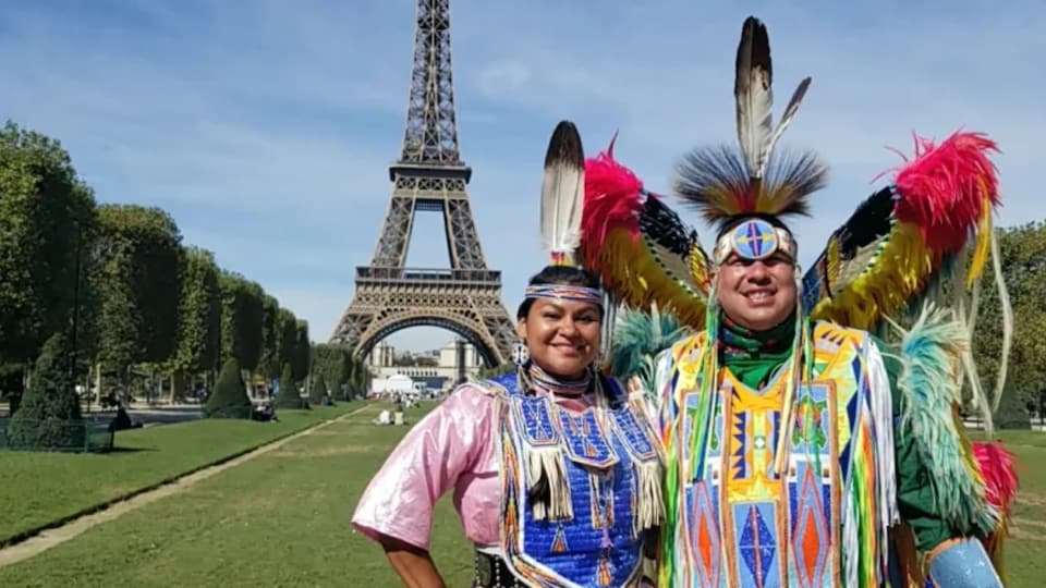 Deux personnes autochtones en tenue traditionnelle posent devant la tour Eiffel.