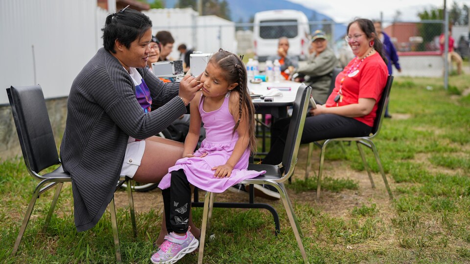 Plusieurs personnes sont attablées à l'extérieur, et une petite fille se fait maquiller par une adulte.