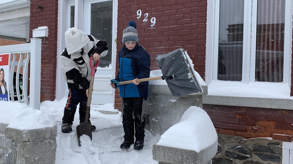 Deux enfants pellettent la neige de leur entrée de maison.