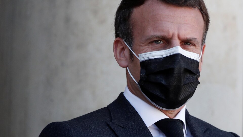 Emmanuel Macron porte un couvre-visage noir et affiche un air sérieux.
