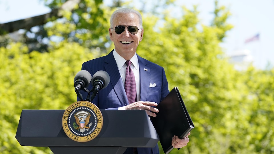 Joe Biden, souriant et portant des lunettes de soleil, tient un porte-documents au lutrin lors d'une conférence de presse à l'extérieur.