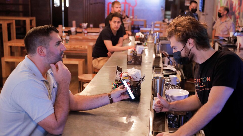 Un serveur derrière le bar regarde le téléphone que lui montre le client assis devant lui.