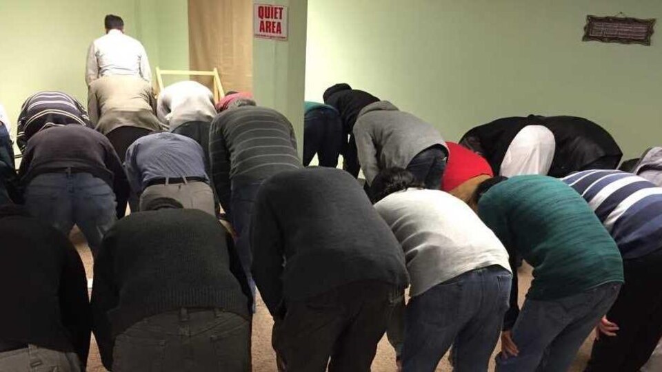 Des hommes, vus de dos, font la prière et se penchant vers l'avant de la salle.