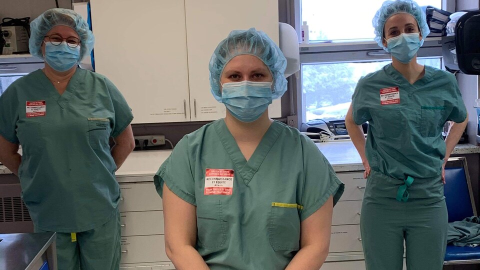 Trois travailleuses en habits d'hôpital avec des masques sur leur bouche et un autocollant où on peut lire "Reconnaissance et équité"