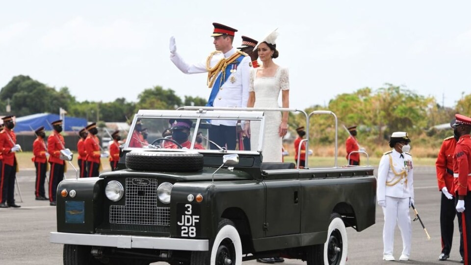 Le prince William et Catherine, debout sur un véhicule, saluent des gens.