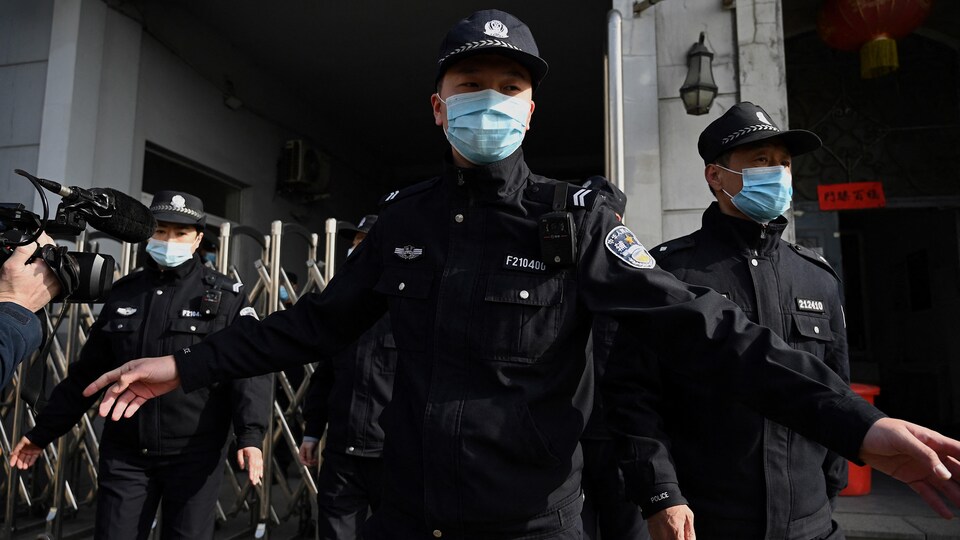 Des policiers chinois font des gestes pour repousser une petite foule, devant une entrée grillagée. 