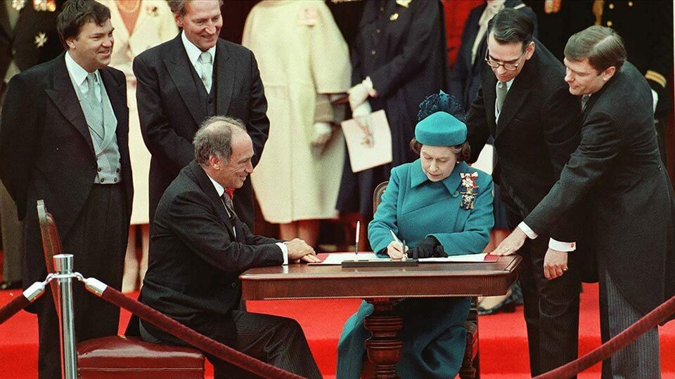 Une femme vêtue de bleu, assise à une table signe un document. Elle est entourée du premier ministre, lui aussi assis près d'elle, et d'autres personnes qui se tiennent debout et sourient, dans un cadre officiel.