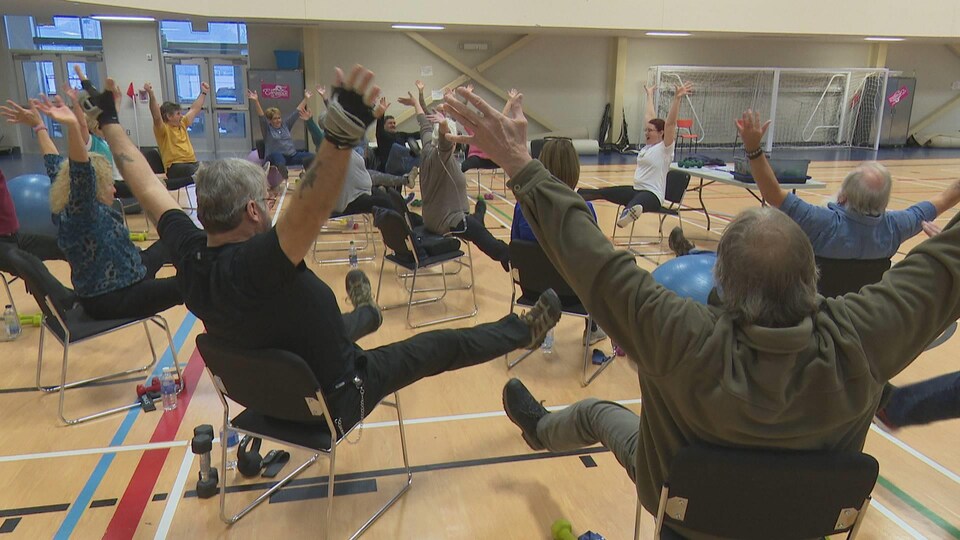 Des hommes et des femmes assis sur des chaises dans un gymnase lèvent les bras et les jambes.
