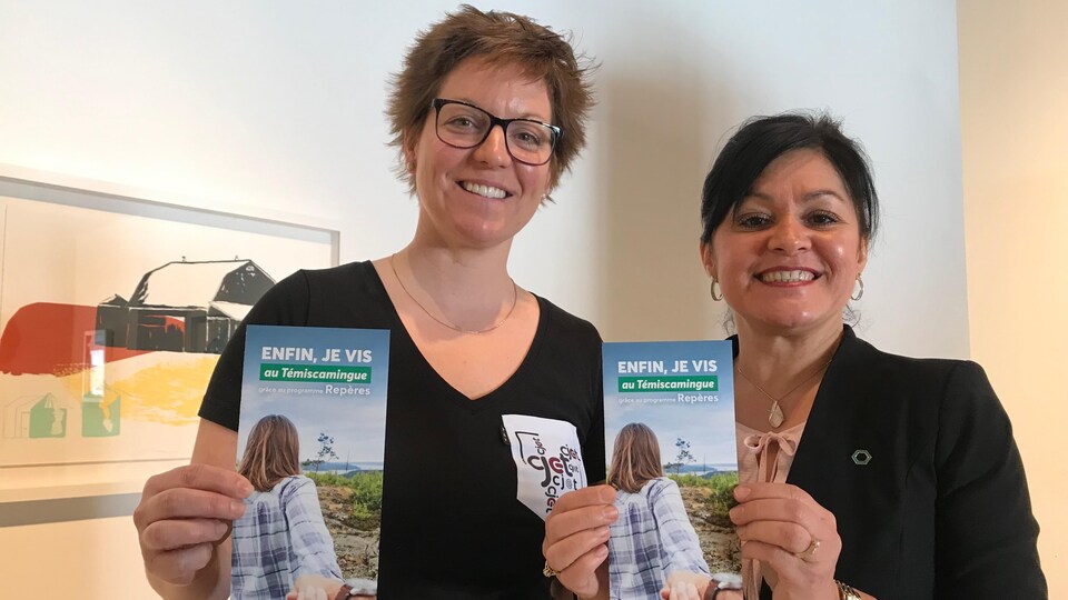 Deux femmes sourient en tenant un dépliant intitulé Enfin, je vis au Témiscamingue.