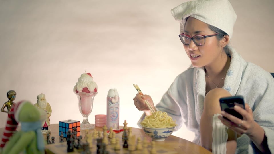 La jeune femme est assise à une table avec un téléphone cellulaire à la main et des baguettes posées dans un bol de nouilles de l'autre. La table est remplie de divers objets, dont un jeu d'échec, un sundae, un cube Rubik et de la mousse à cheveux.