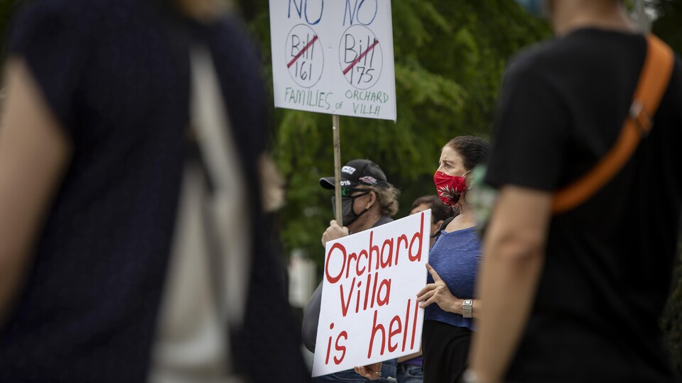 Une femme masquée tient une pancarte sur laquelle on peut lire "Orchard Villa is hell". 
