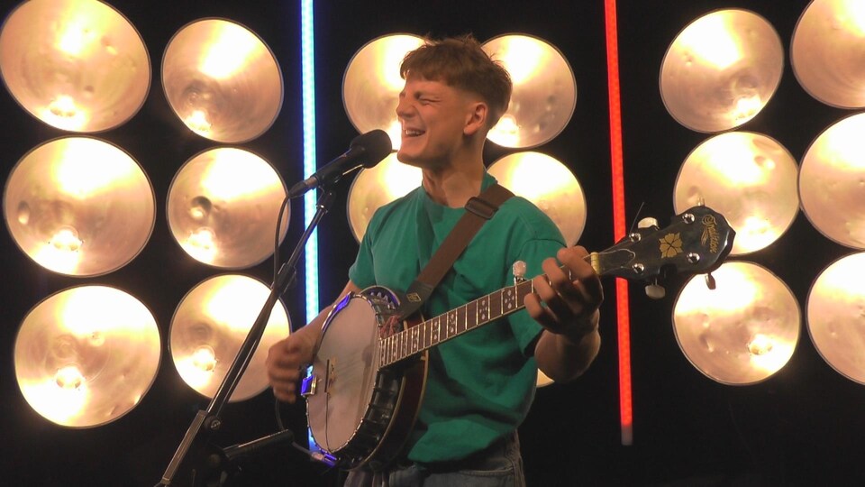 Le jeune homme chante dans un micro sur pied et s'accompagne au banjo sur un plateau de télévision.