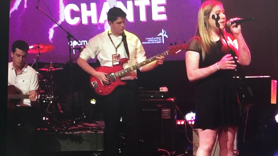 Une jeune femme chante en tenant un micro à sa bouche tandis qu'un jeune homme joue de la guitare derrière elle.