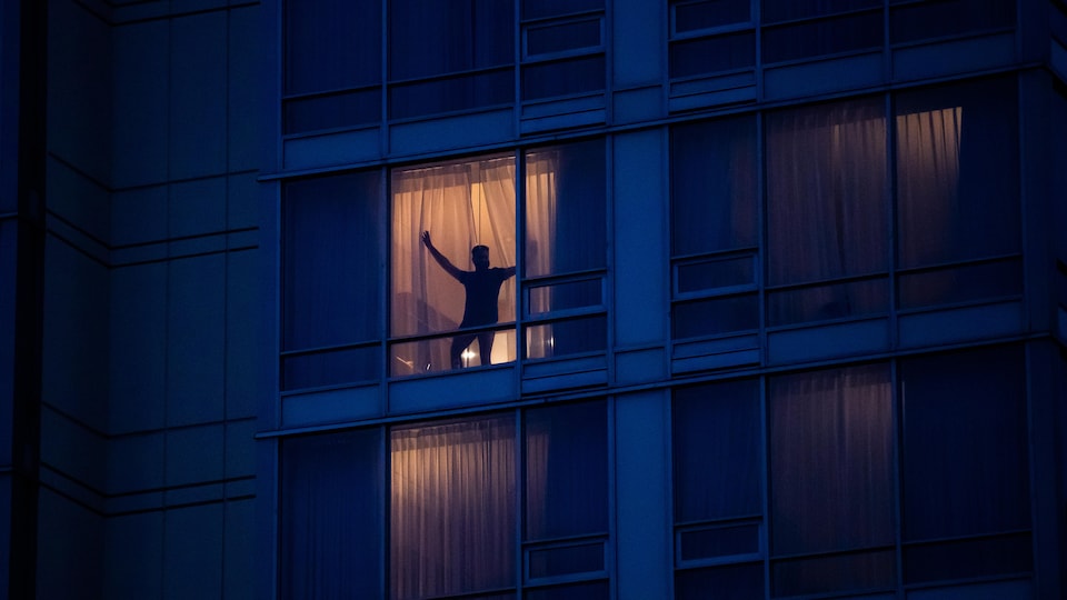 Photographiée de l'extérieur, silhouette d'un homme qui ouvre les rideaux de sa chambre d'hôtel, le soir.
