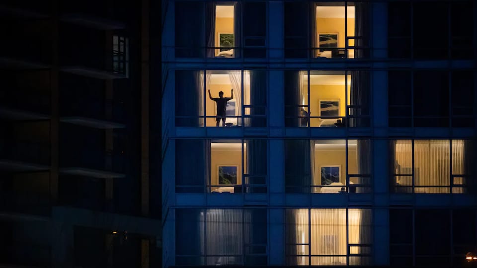 Un homme ouvre les rideaux de sa chambre d'hôtel après le coucher du soleil.