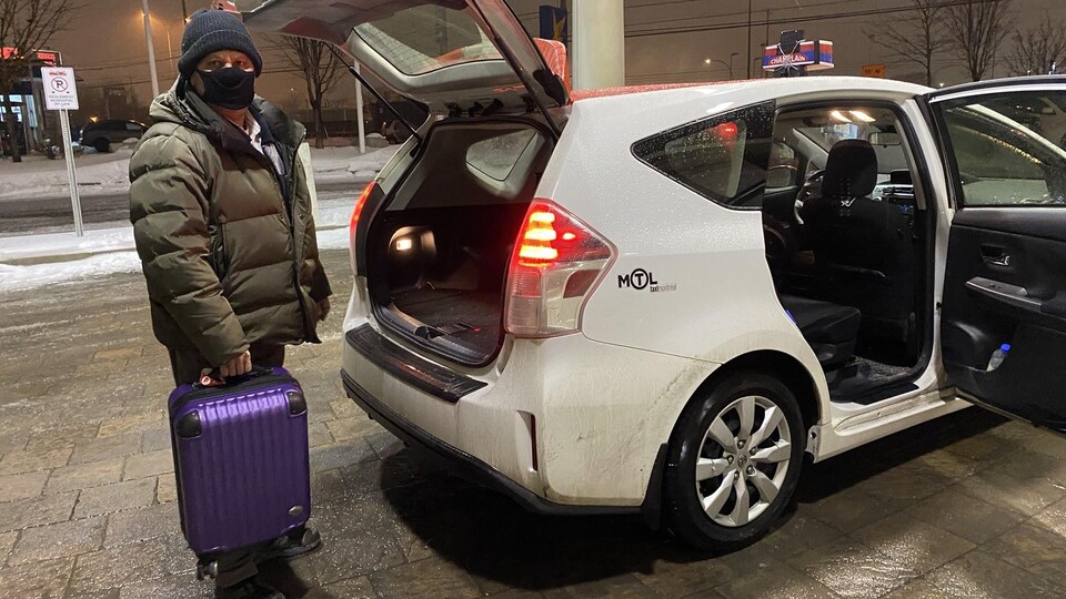 Chauffeur de taxi portant un masque, une valise mauve dans ses mains et regardant la caméra