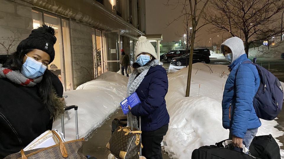 Trois personnes portant tuques, manteaux et masques et transportant leurs valises regardent la caméra.