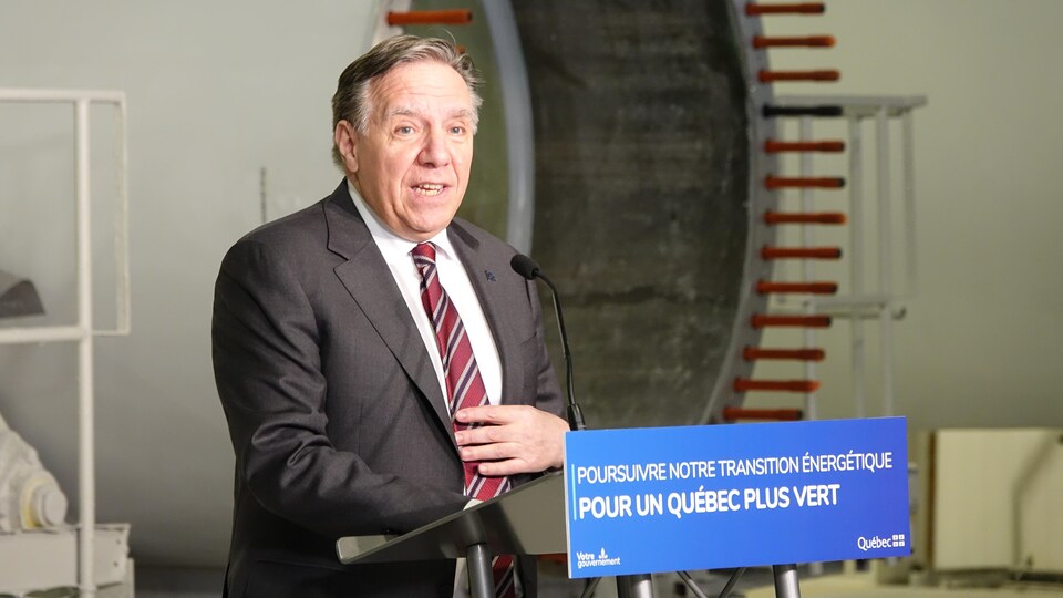 Le premier ministre du Québec tient une conférence de presse. Sur son pupitre est inscrit : « Poursuivre notre transition énergétique pour un Québec plus vert». Derrière lui, une pale d'éolienne. 