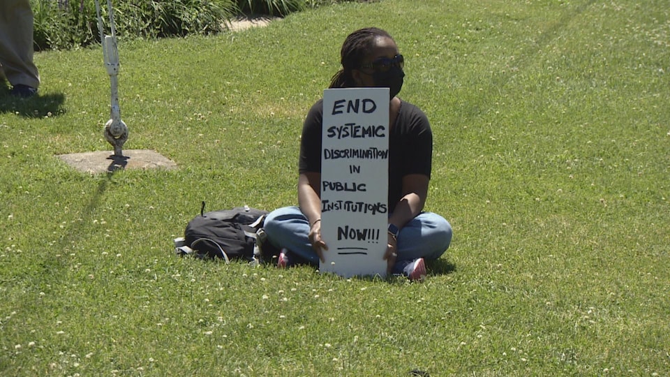 Une manifestante tient une pancarte où il est écrit qu'il faut faire cesser la discrimination systémique au sein des institutions publiques.