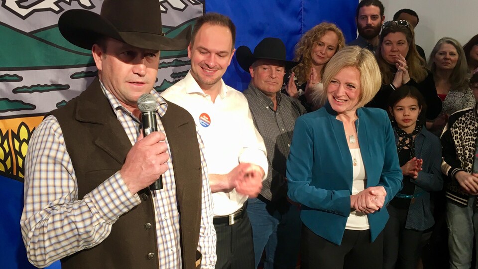 Un homme avec un chapeau cowboy tient un micro. À ses côtés, un homme et une femme sourient. Derrière se trouve une foule de personnes ainsi que le drapeau de l'Alberta.