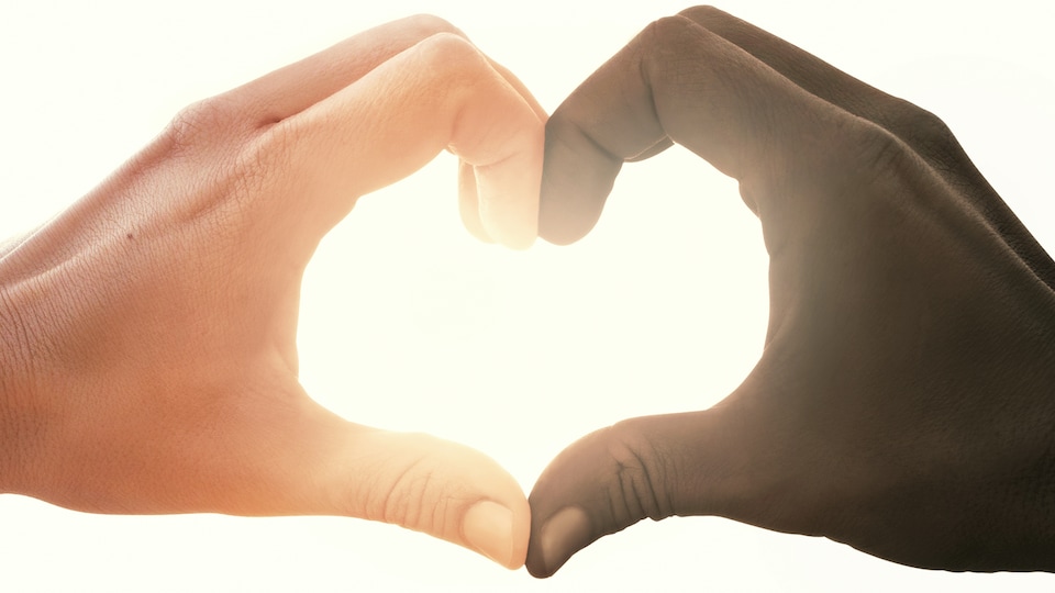 Deux mains jointes en forme de coeur, une d'une personne noire et l'autre d'une personne blanche. La lumière filtre au travers du coeur qui est ainsi formé. 