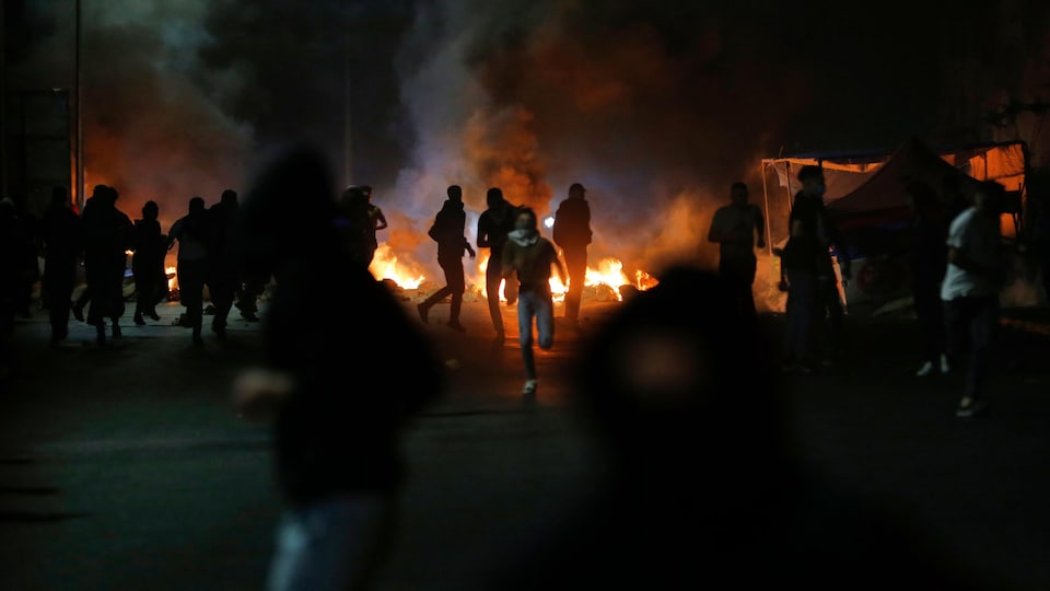 Des Palestiniens se sauvent des soldats israéliens dans une rue sombre tandis que des feux sont allumés au loin.