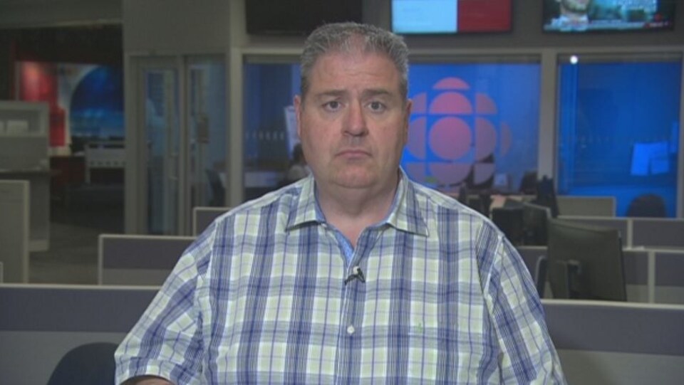 Un homme en chemise à carreaux devant les écrans d'ordinateur d'une salle de nouvelles aux couleurs de Radio-Canada.